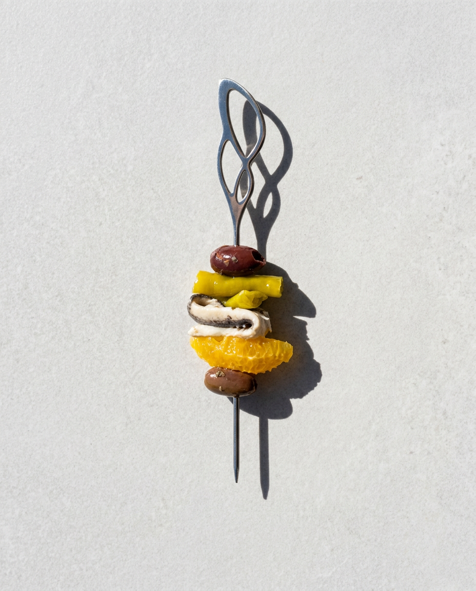 Presentación vertical de una gilda gourmet de boquerón, naranja y aceituna negra ensartado en brocheta metálica con diseño de curvas