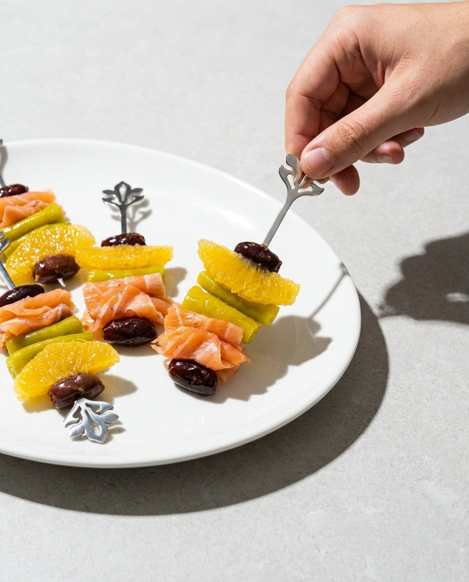 Mano sirviendo un pincho de salmón y naranja con una brocheta metálica decorativa de flor en un plato