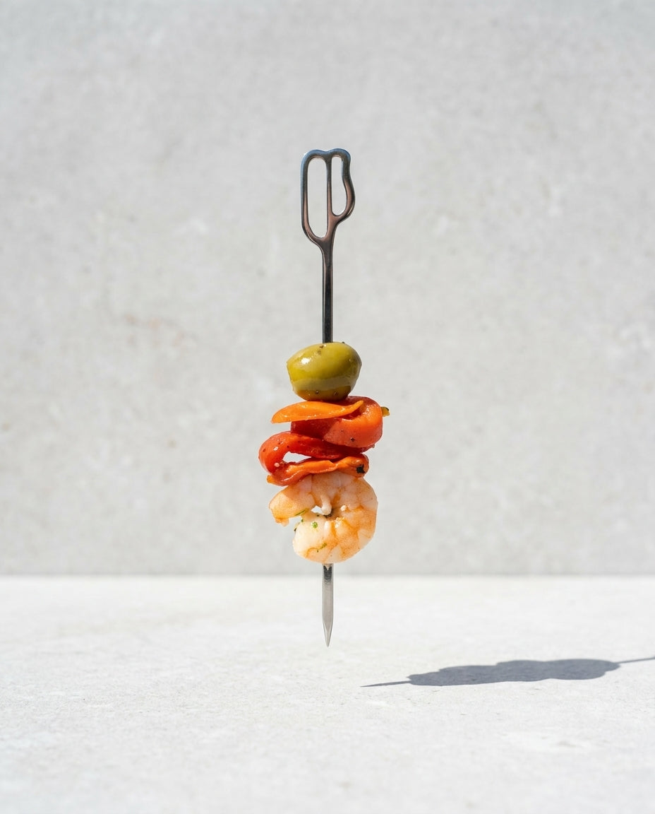 Presentación vertical de un pincho de gilda con gamba, pimiento y aceituna verde ensartado en una brocheta metálica de diseño inspirada en la Casa Batlló